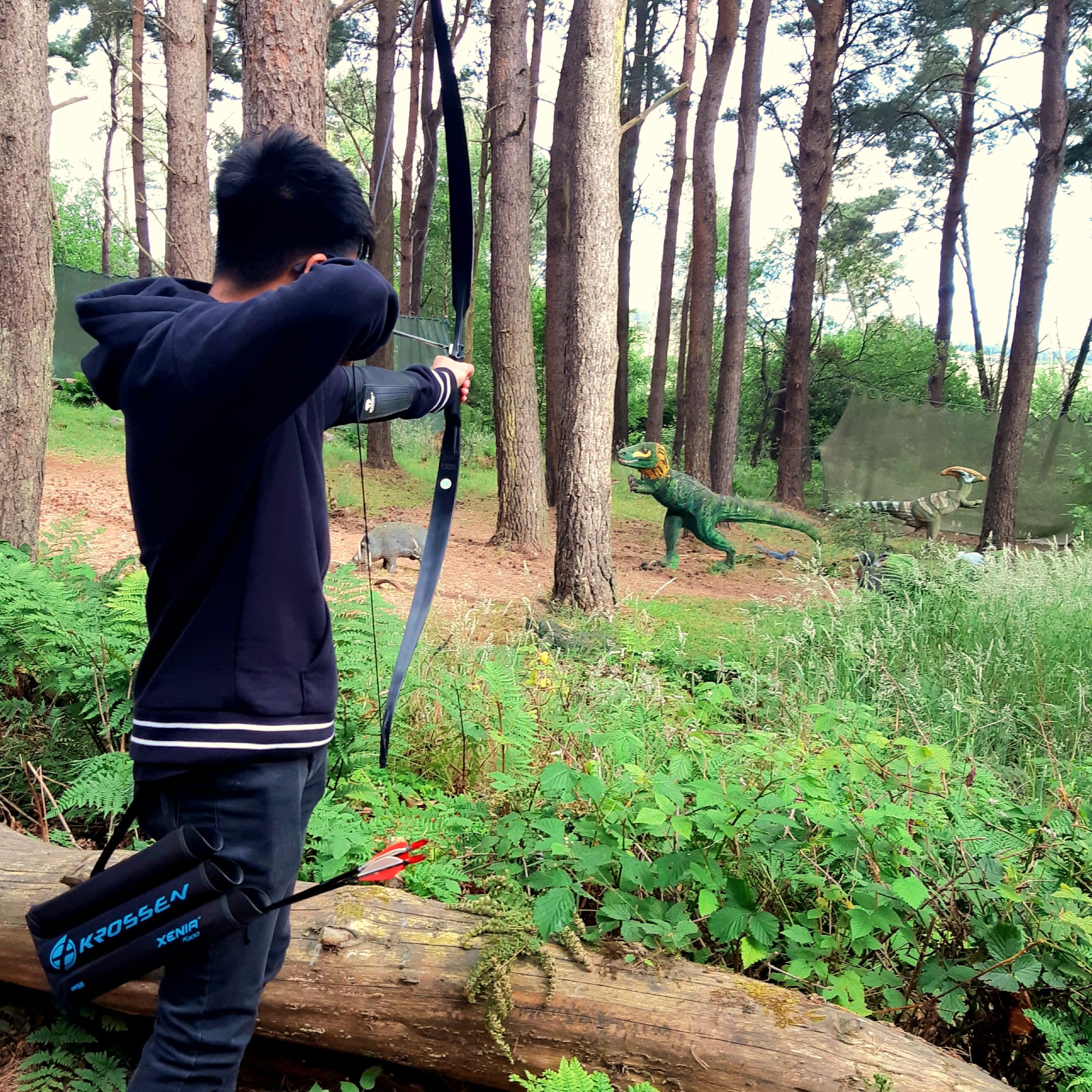 young person stood at full draw, aiming arrow at dinosaur in the background